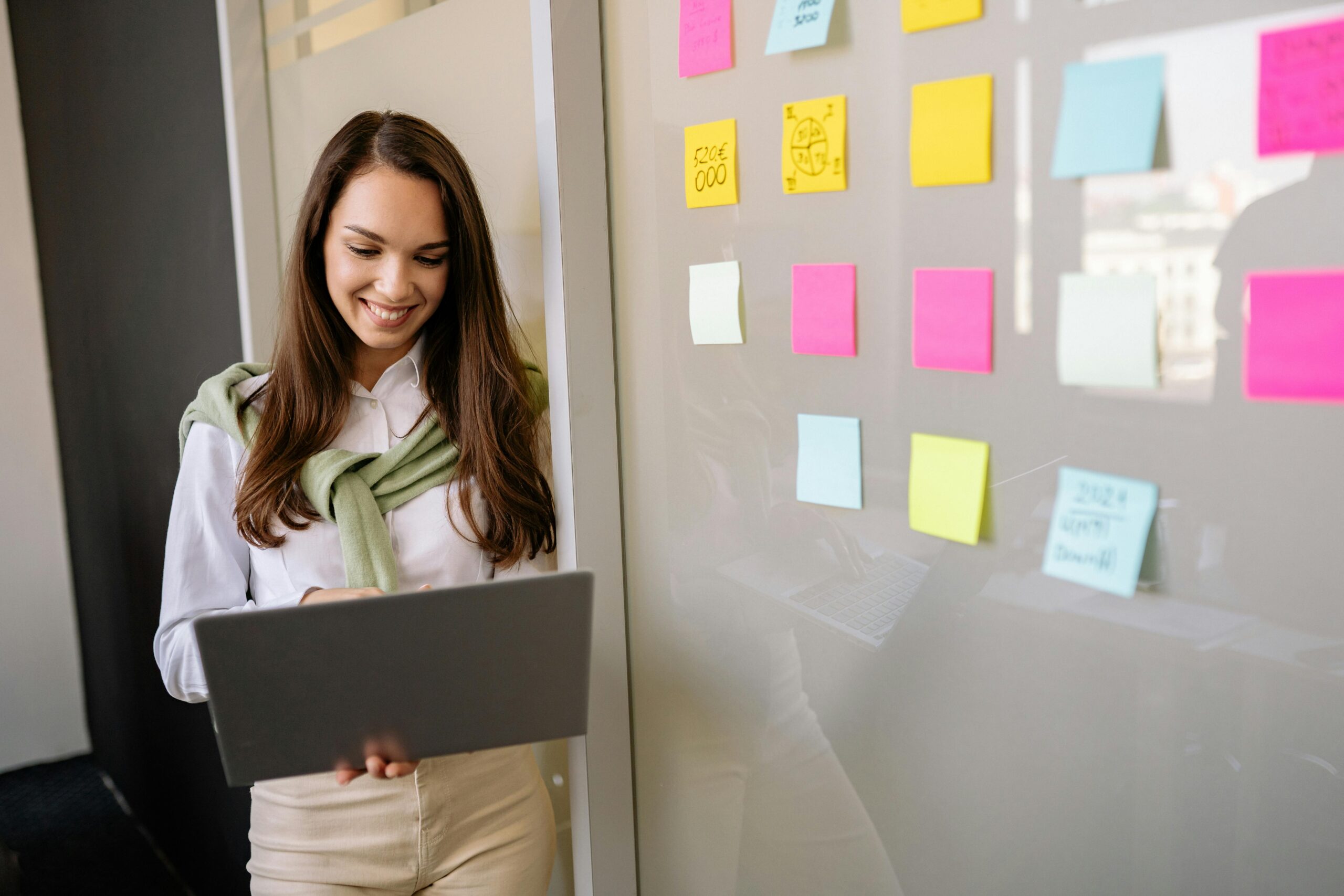 Smiling woman uses laptop in office, planning with colorful sticky notes on glass wall.